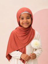 Girl wearing the rust jersey hijab for girls, holding a white dahlia, front view