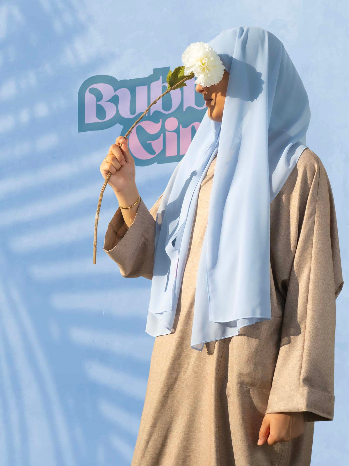 Person wearing a light blue hijab and beige outfit holding a white flower against a blue background with 'Bubu Girl' branding.