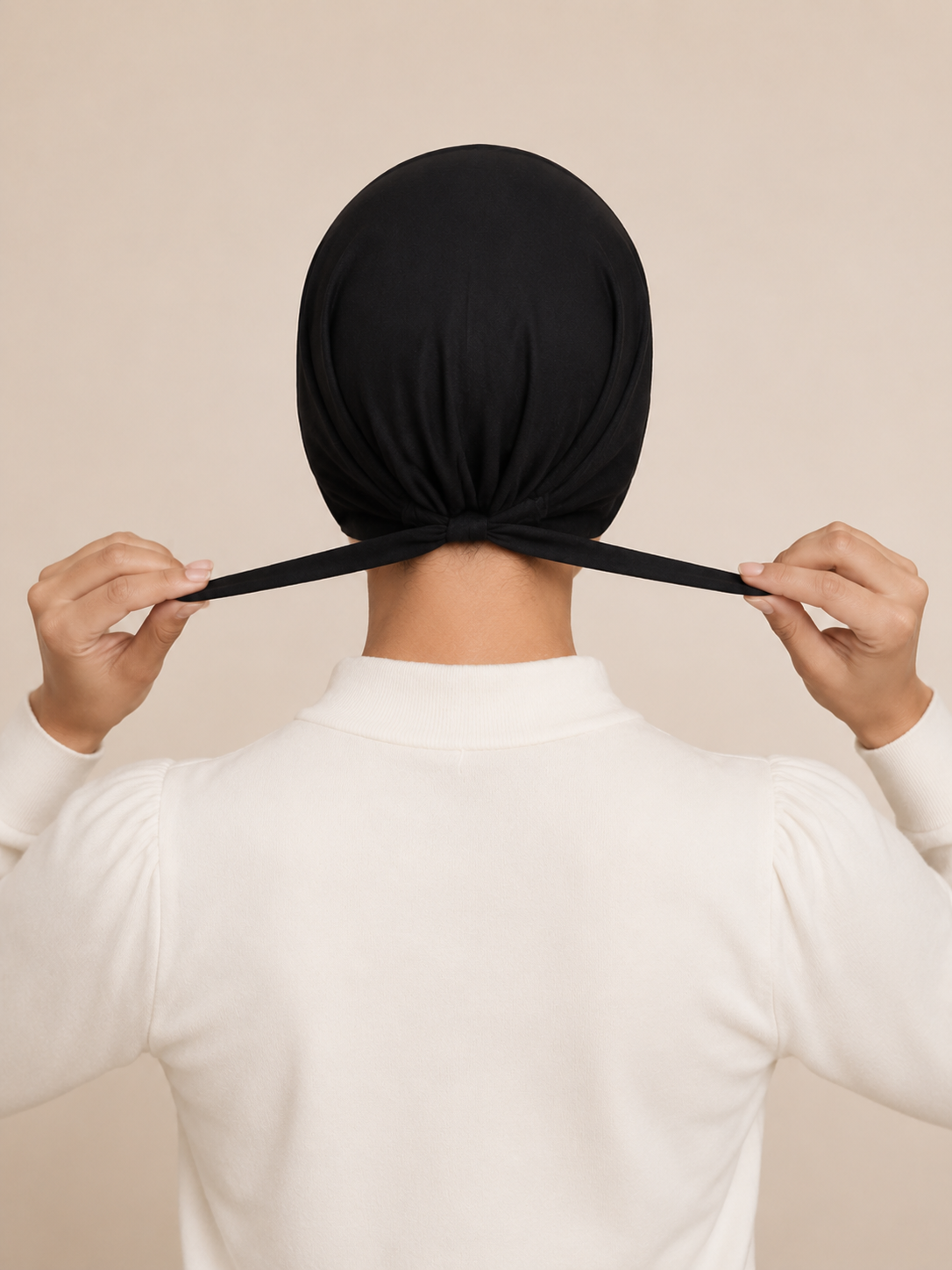 A person viewed from behind wearing a black full-head fabric cap or bonnet and pulling the elastic band across the back of the head, against a plain warm beige background.