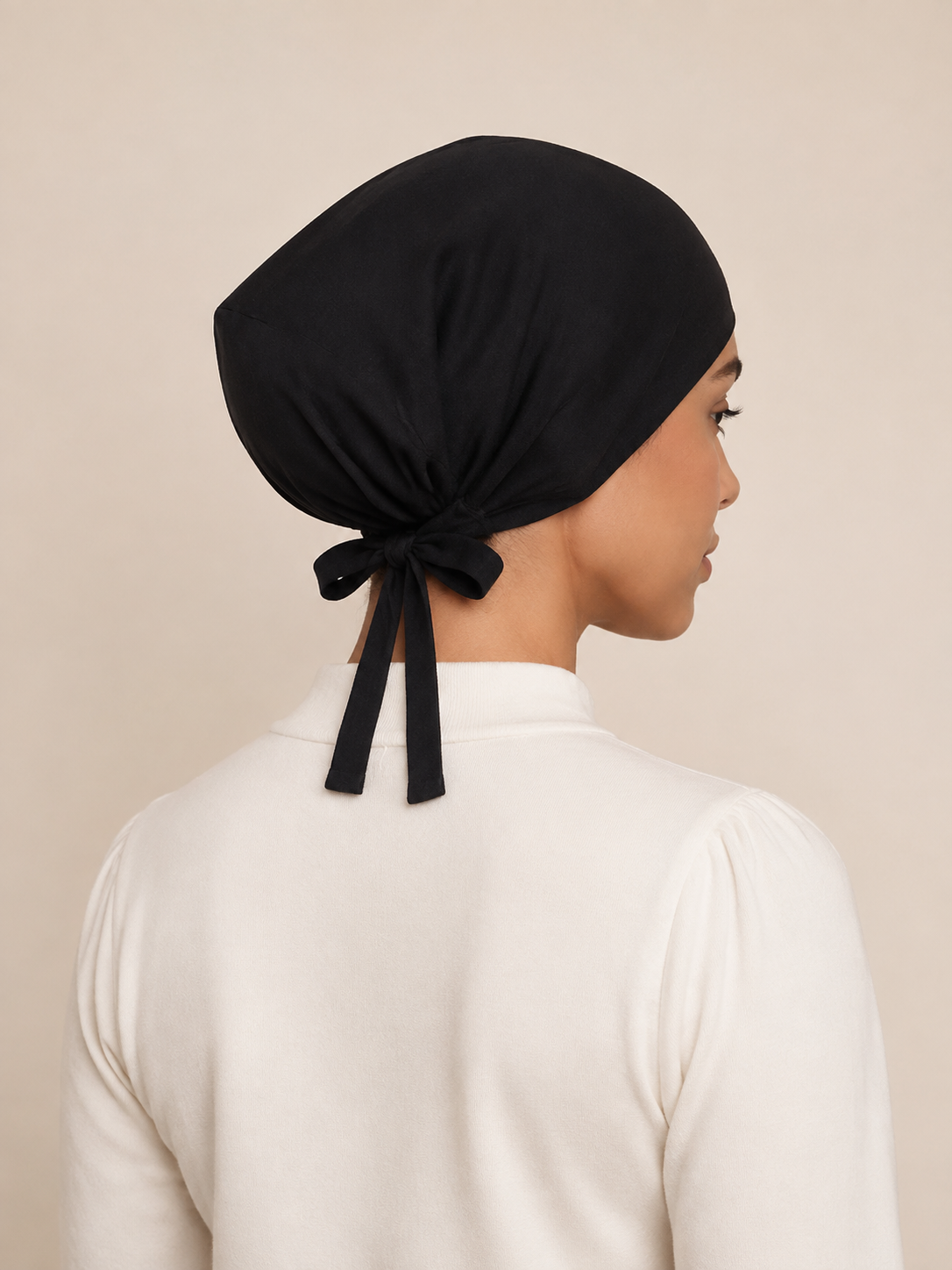 A woman shown from the back wearing a black fabric head covering tied at the back, paired with a white long-sleeve top against a plain beige background.