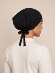 A woman shown from the back wearing a black fabric head covering tied at the back, paired with a white long-sleeve top against a plain beige background.