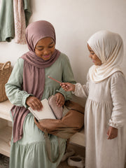 Two girls at a hallway bench, one in the dusty rose jersey hijab for girls with a satchel