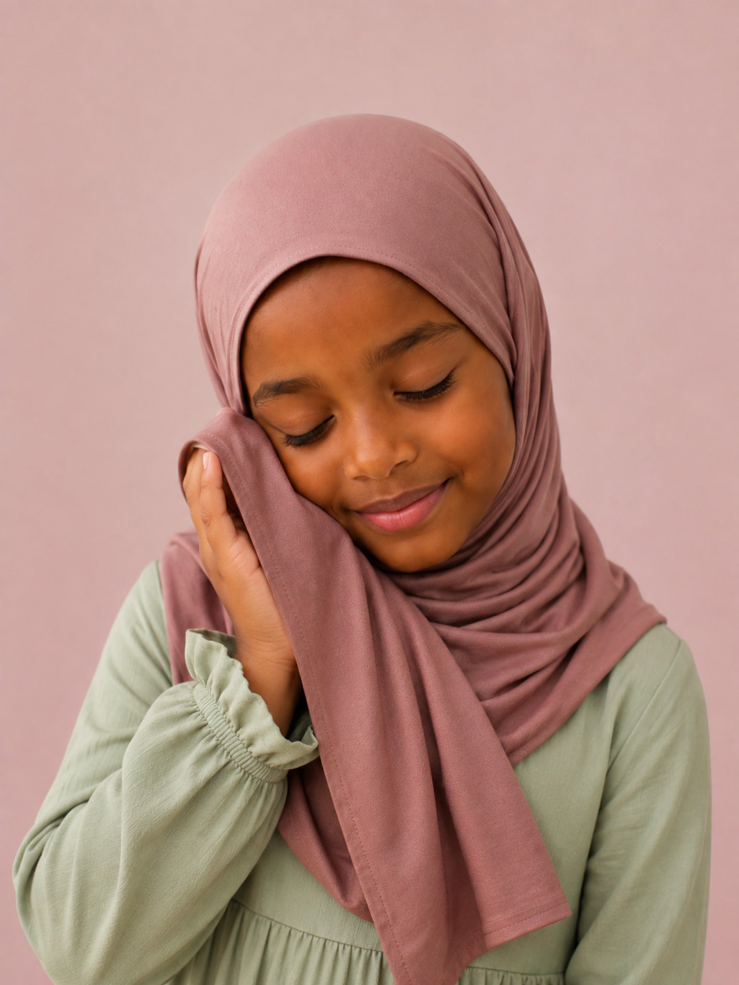 Girl holding the dusty rose jersey hijab for girls against her cheek, eyes closed