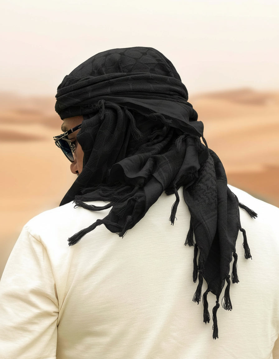 Man in a desert wearing an all black keffiyeh wrapped over the head and across the face, tassels hanging at the shoulder