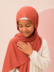 Girl resting a hand against the rust jersey hijab for girls, eyes lowered