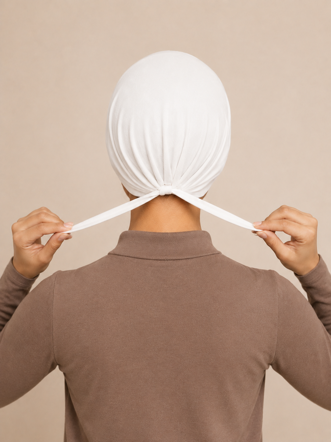 A person viewed from behind wearing a white fabric head covering and adjusting the ties with both hands, against a plain beige background.