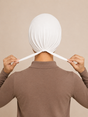 A person viewed from behind wearing a white fabric head covering and adjusting the ties with both hands, against a plain beige background.