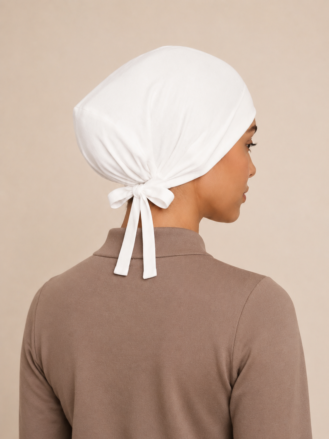 A woman shown from the back wearing a white fabric head covering tied in a bow at the back, paired with a taupe-brown long-sleeve top against a plain beige background.